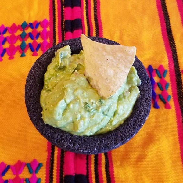 Guacamole in a dark stone molcajete with a tortilla chip, on a colorful Mexican serape.
