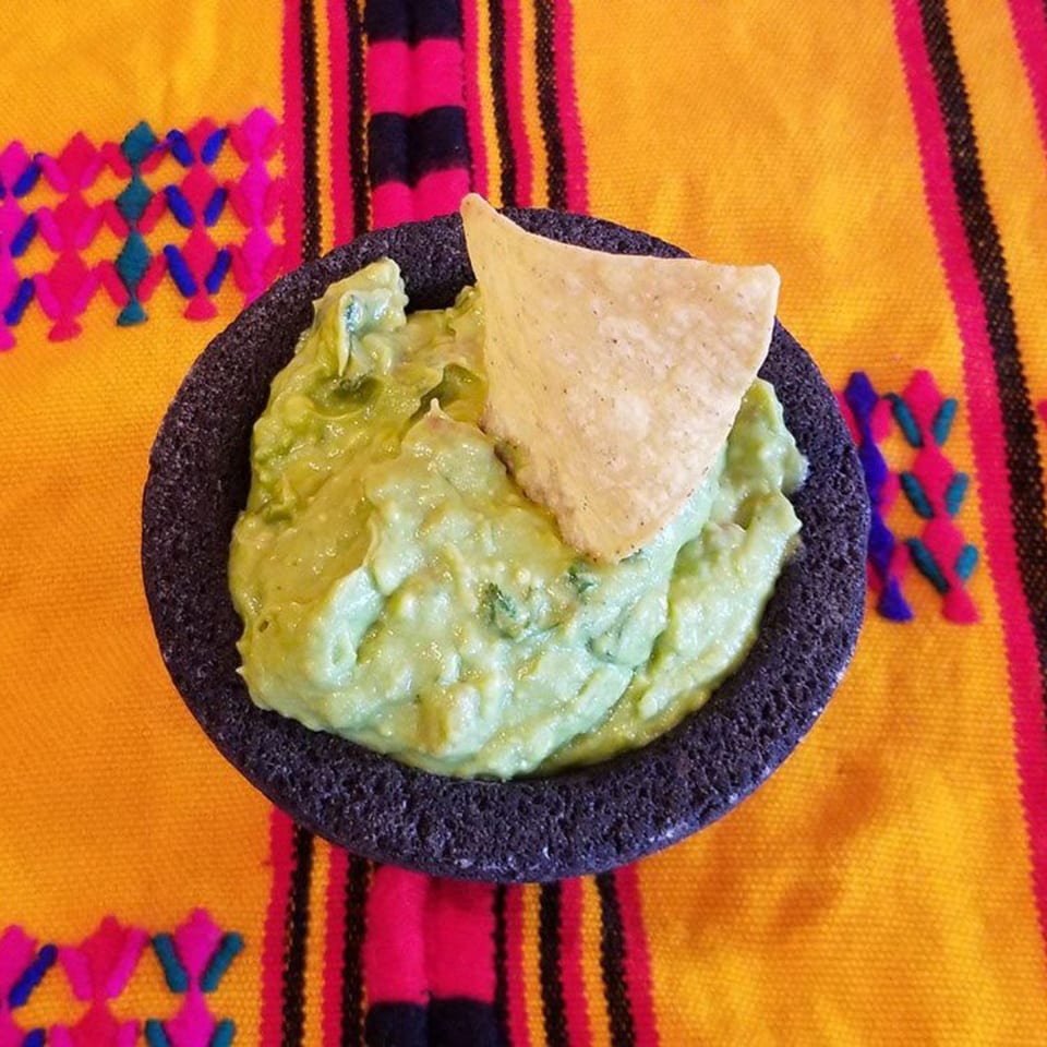 Guacamole in a dark stone molcajete with a tortilla chip, on a colorful Mexican serape.