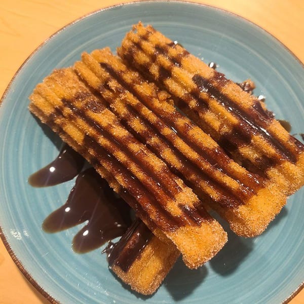 Golden churros drizzled with chocolate sauce and sprinkled with sugar on a blue plate.