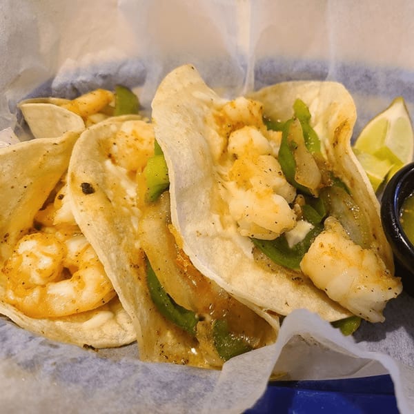 Three shrimp tacos with green peppers and onions, served with lime wedges and green salsa.
